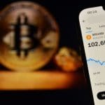 Das Bild eines Mobiltelefons zeigt den Bitcoin-Preis über 100.000 $, mit einer BTC-Münze im Hintergrund