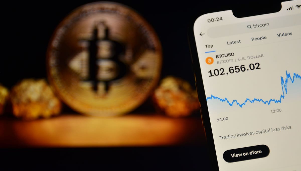 Das Bild eines Mobiltelefons zeigt den Bitcoin-Preis über 100.000 $, mit einer BTC-Münze im Hintergrund